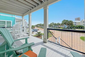 A covered outdoor area with green chairs and a table, overlooking a paved area with palm trees and buildings.