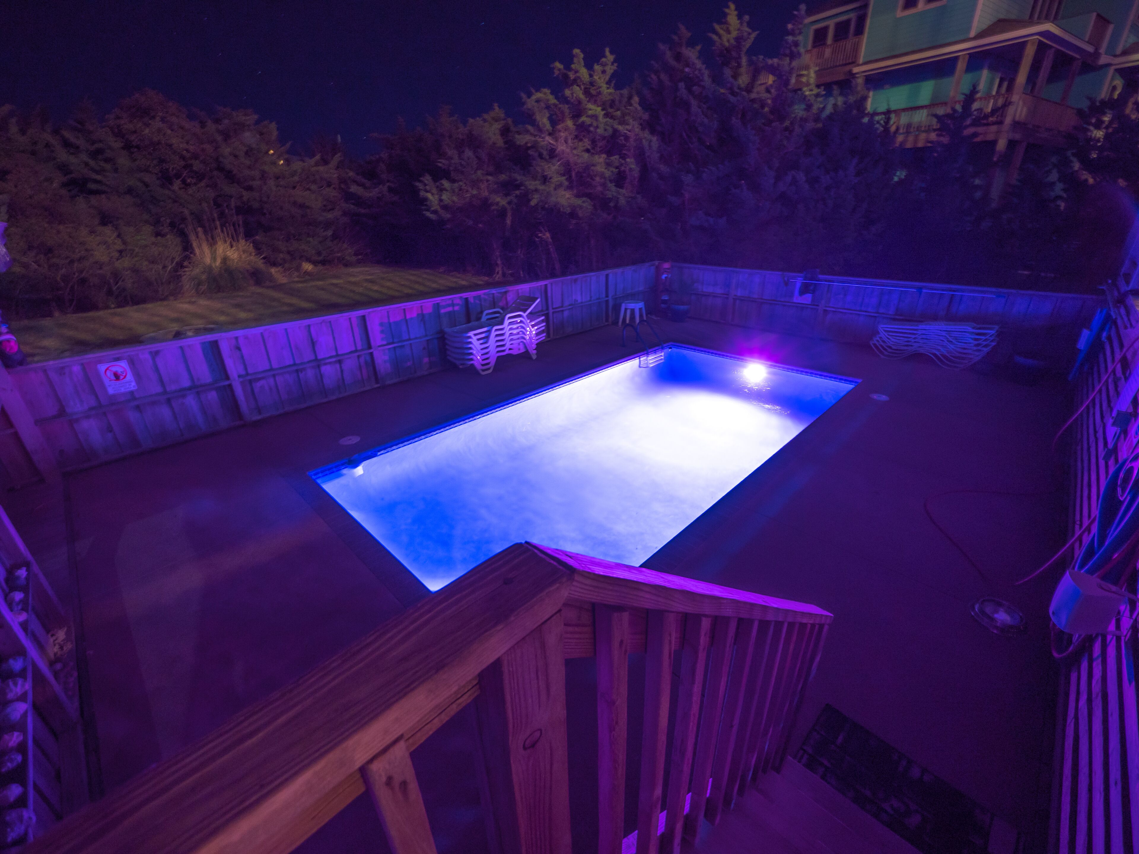 A rectangular swimming pool with a wooden deck surrounding it, set against a backdrop of trees and a building.