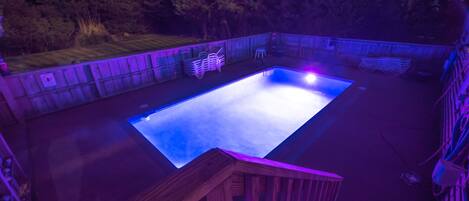 A rectangular swimming pool with a wooden deck surrounding it, set against a backdrop of trees and a building.