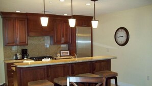A kitchen with wooden cabinets, a stainless steel refrigerator, pendant lights, a clock, and a marble countertop.