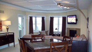 A dining area with a glass-top table and wooden chairs, a sofa, and a television mounted on the wall.