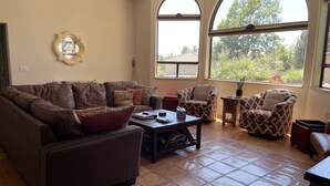 A living room with a brown leather sofa, two patterned armchairs, a coffee table, and large arched windows offering a view of greenery. - Casa de Saltillo - single level home, gorgeous views, close to trails. (Sedona)