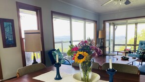 Un comedor con mesa de madera, sillas, un jarrón con flores y vistas amplias a través de grandes ventanales.