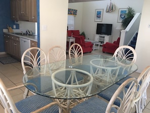 Un coin repas avec une table en verre et des chaises blanches, une cuisine avec des meubles en bois, un canapé rouge, un téléviseur et un escalier.