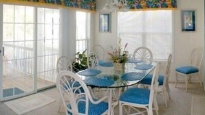A dining area with a glass-top table surrounded by white chairs with blue cushions. There is a window with blinds and a colorful mural on the wall above the window. - Professionally Decorated,100 Yards to Beach, Shared Pool (Surfside Beach)
