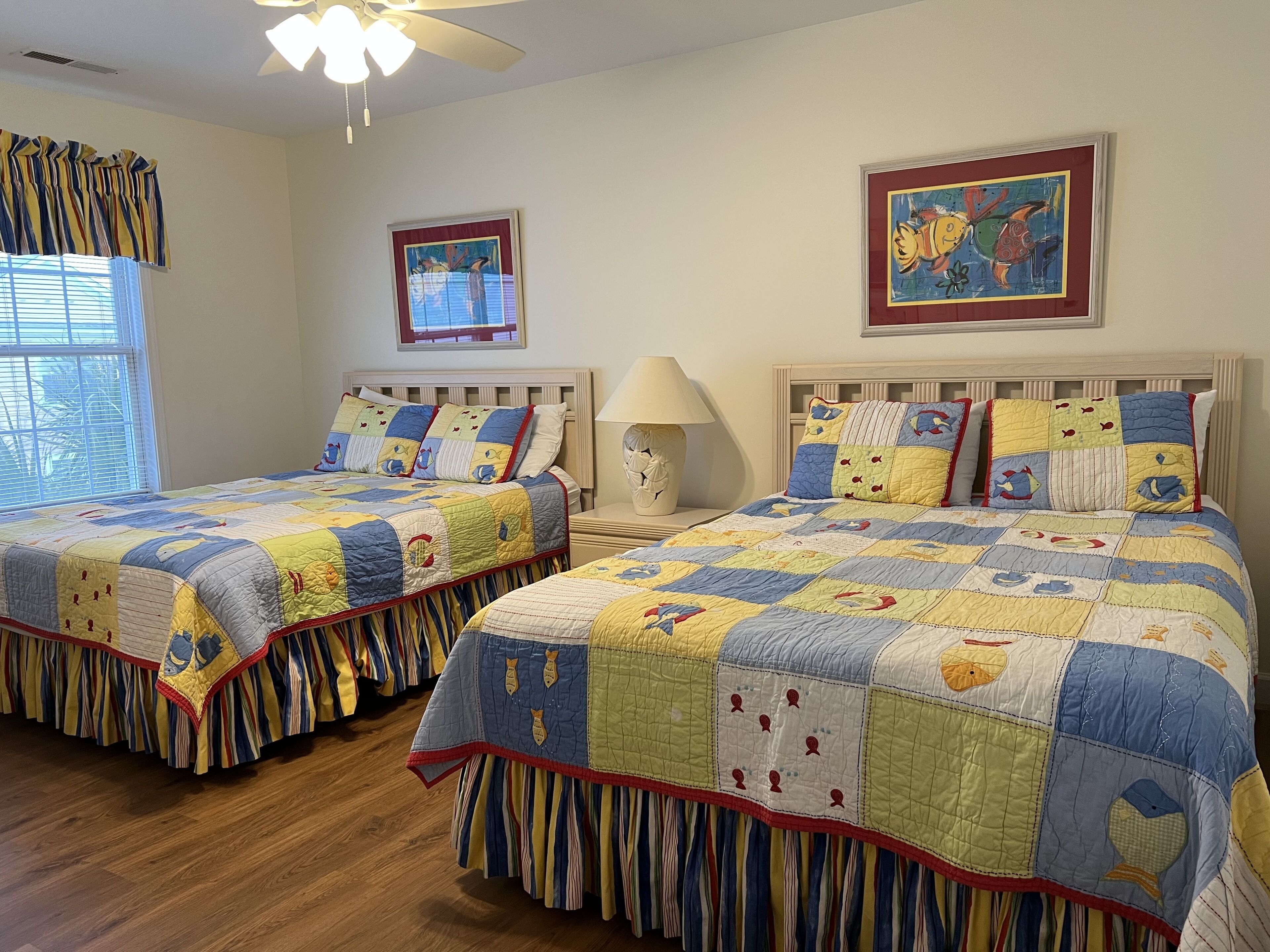Un dormitorio con dos camas, cada una con colcha acolchada con motivos de peces. Hay una ventana con cortinas, un ventilador de techo y cuadros en las paredes.