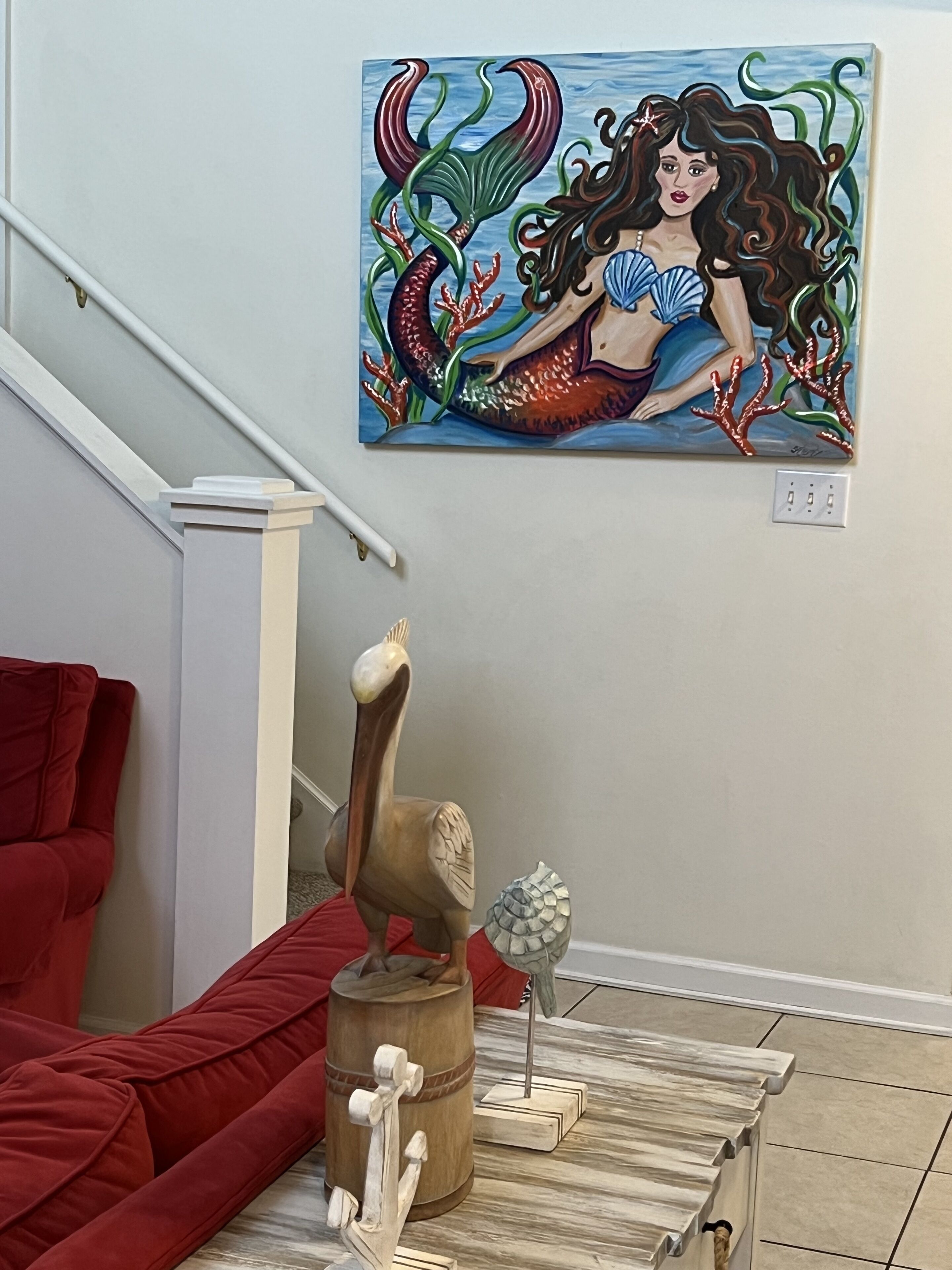 A living room with a red sofa, a wooden coffee table with nautical-themed decor, and a painting of a mermaid on the wall.