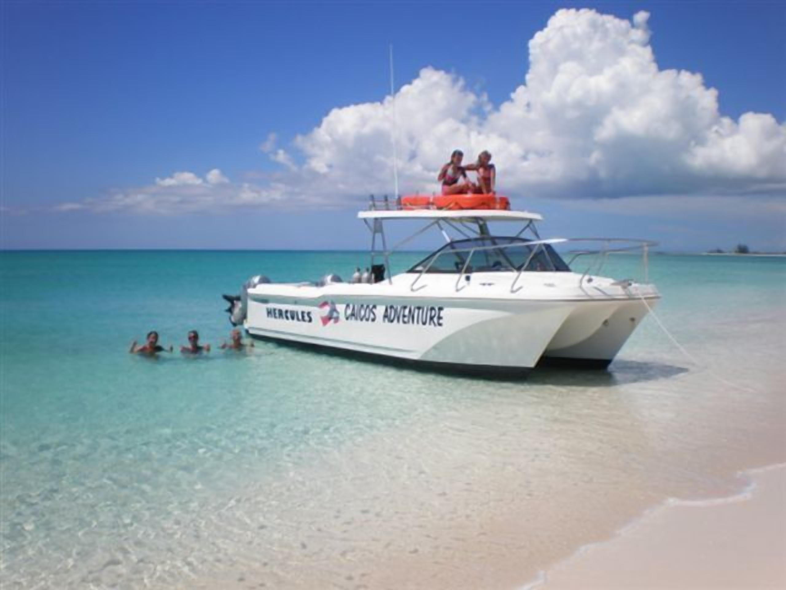 Una lancha llamada 'Hercules' con la leyenda 'Caicos Adventure' en su lateral, fondeada en una playa de aguas turquesas cristalinas y orilla arenosa.