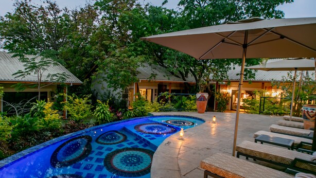 Une piscine dont le carrelage présente un motif géométrique, entourée de chaises longues et de parasols, avec un bâtiment et une végétation luxuriante en arrière-plan.
