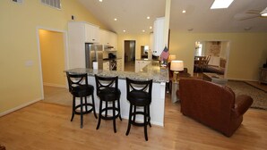 A modern kitchen with a central island, bar stools, and an open-plan living area with a couch and a dining table. - A Second Wind! With Sliding board/diving board!! (Huddleston)
