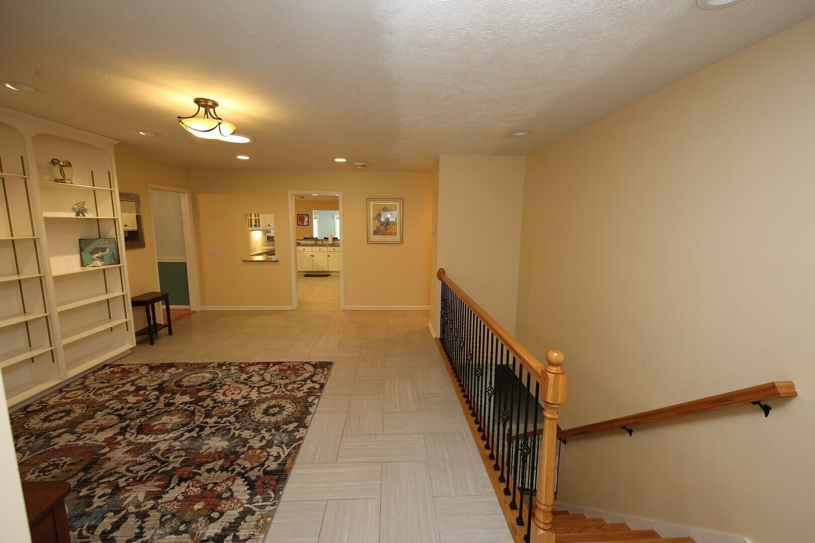 A spacious hallway with a patterned rug, a wooden staircase with a black railing, and a kitchen area visible through an open door.
