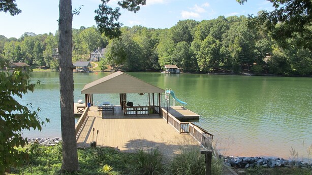 A wooden dock with a covered seating area, a playground slide, and a view of a lake surrounded by trees. - A Second Wind! With Sliding board/diving board!! (Huddleston)