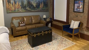 A living room with a brown leather couch, a blue armchair, a black trunk, a large painting of a forest, and a framed poster on the wall. - Studio Loft in historic Silverton Building (Silverton)