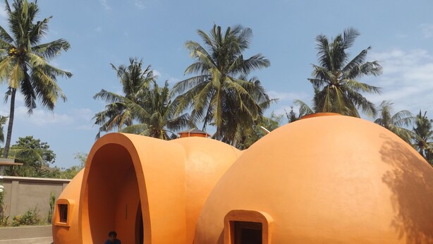 A structure with two large, orange, dome-shaped rooms with small windows, a person sitting inside one room, and palm trees in the background. - Pumpkin Villa  Lombok (Senggigi)