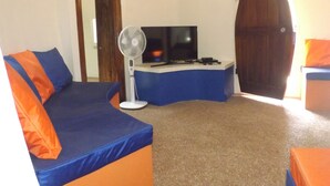 A room with a curved blue sofa, a standing fan, a flat-screen TV, and a wooden door. - Pumpkin Villa  Lombok (Senggigi)