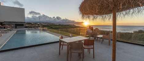 A rooftop pool area with a view of the sunset over the ocean, a dining table, and chairs.