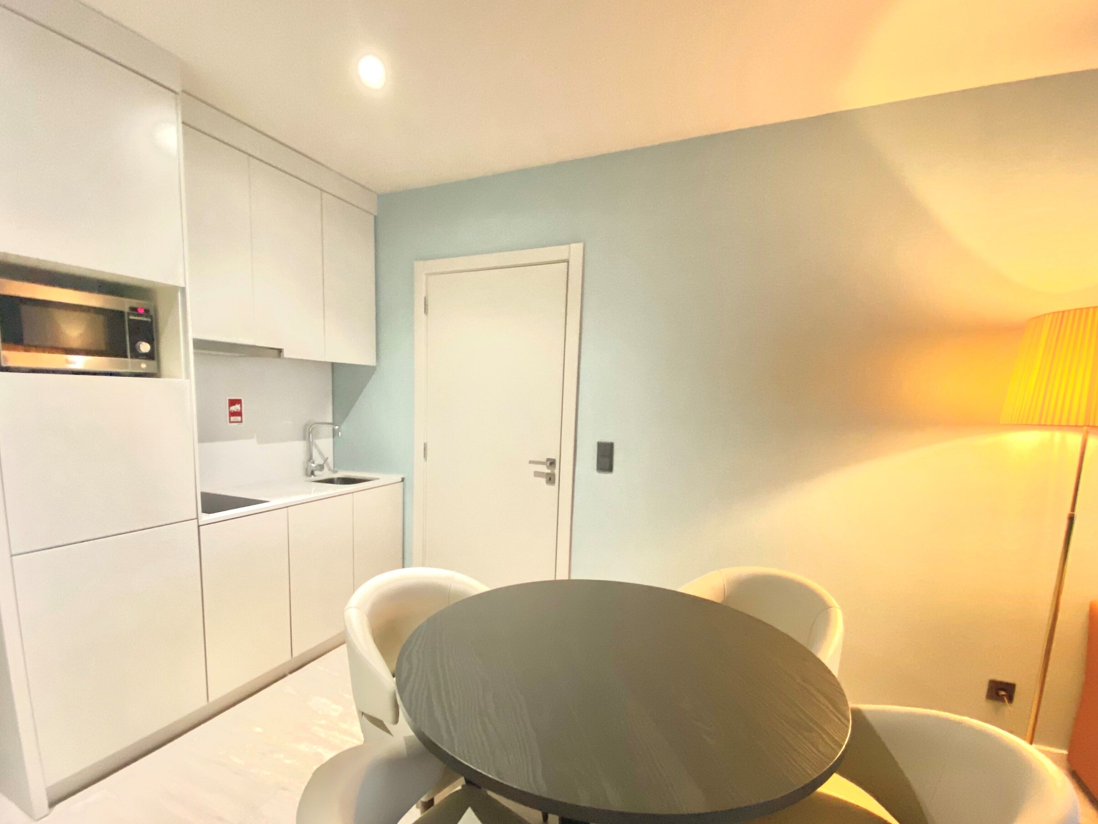 Een kleine keuken met witte kastjes, een magnetron en een spoelbak. Er staat een rond tafel met vier stoelen en een deur op de achtergrond.
