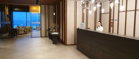 Área de recepção de hotel com design moderno, incluindo um balcão de atendimento, área de estar com sofás, uma grande janela com vista para o oceano e uma planta decorativa.