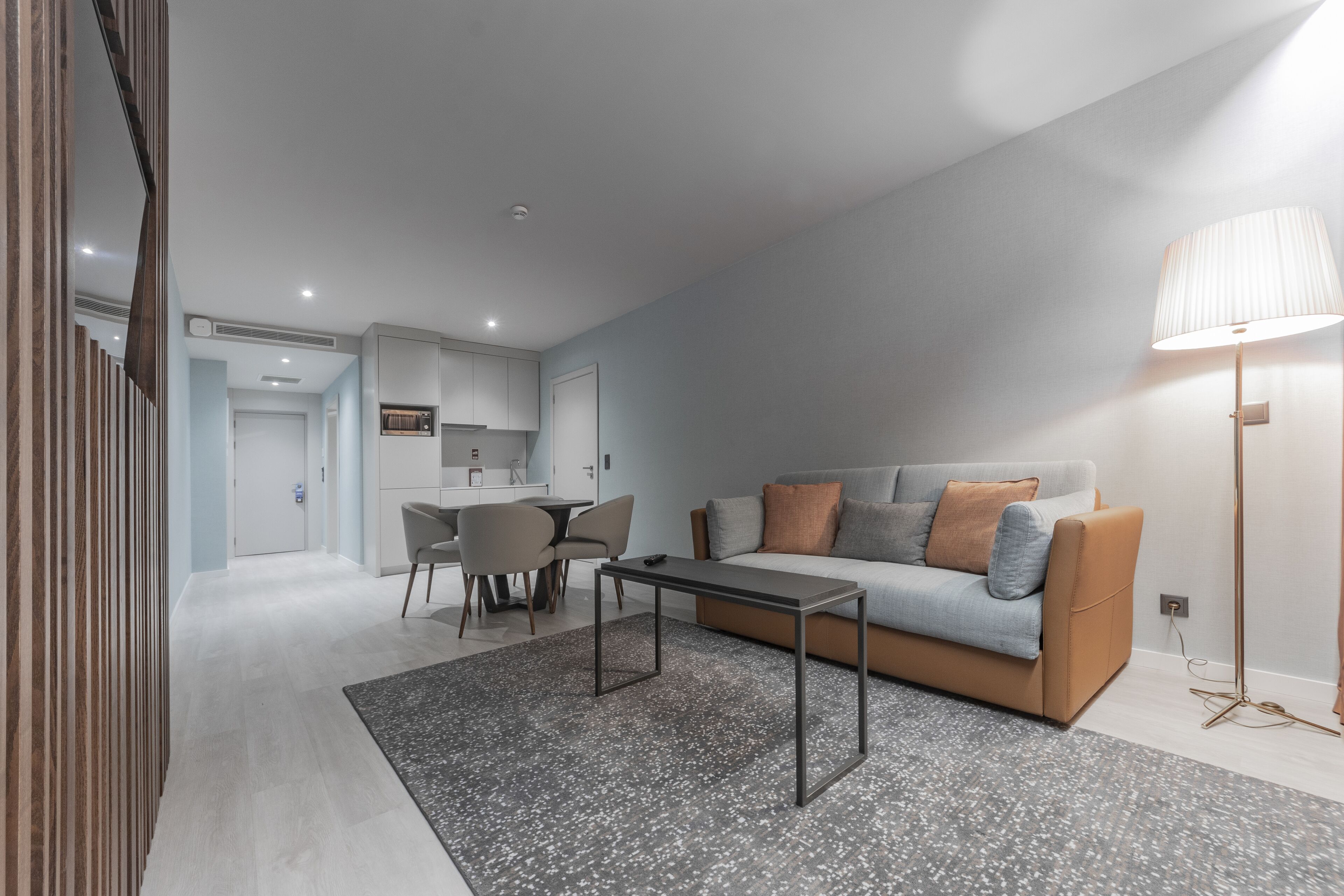 Una sala de estar moderna con un sofá, una mesa de centro y una lámpara. Hay un comedor con sillas y una mesa. El ambiente tiene piso y paredes de tonos claros.