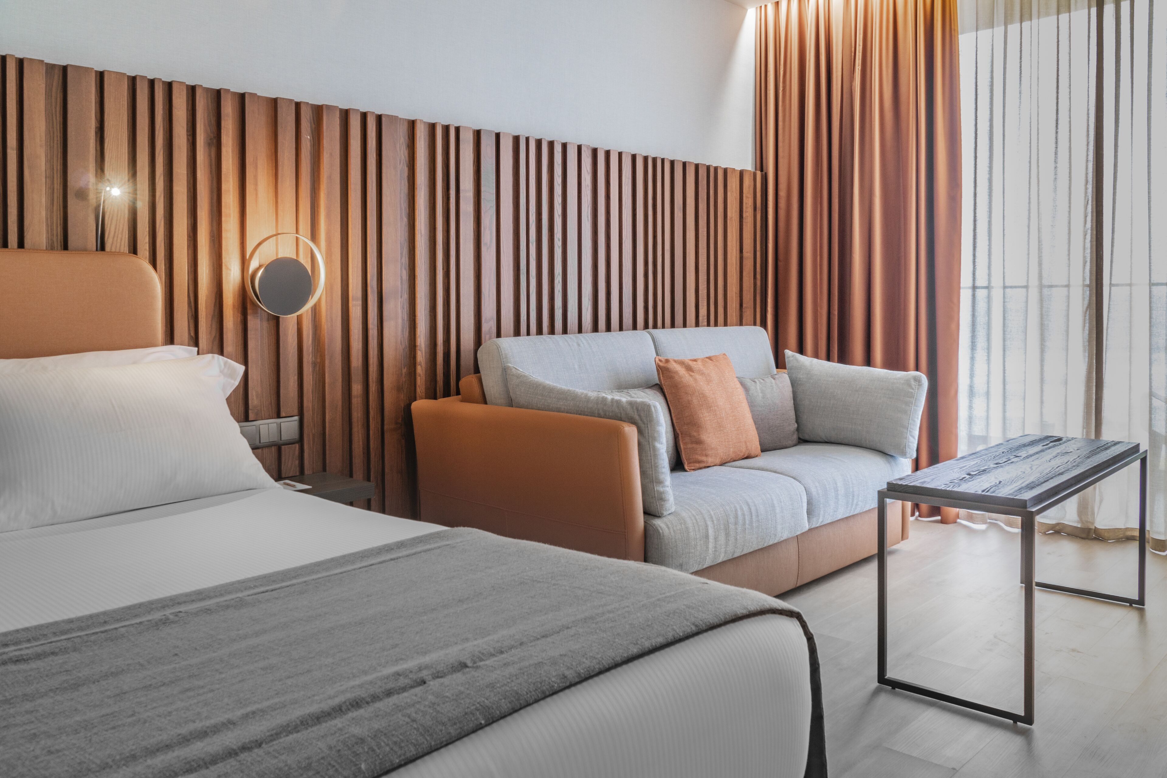 A modern hotel room with a large bed, a sofa, a small table, and a wooden accent wall.