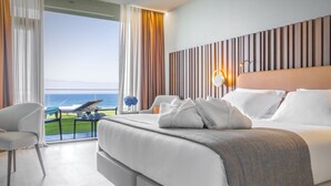 Een moderne hotelkamer met een groot bed, uitzicht op zee, en een balkon met een tafel en stoelen.