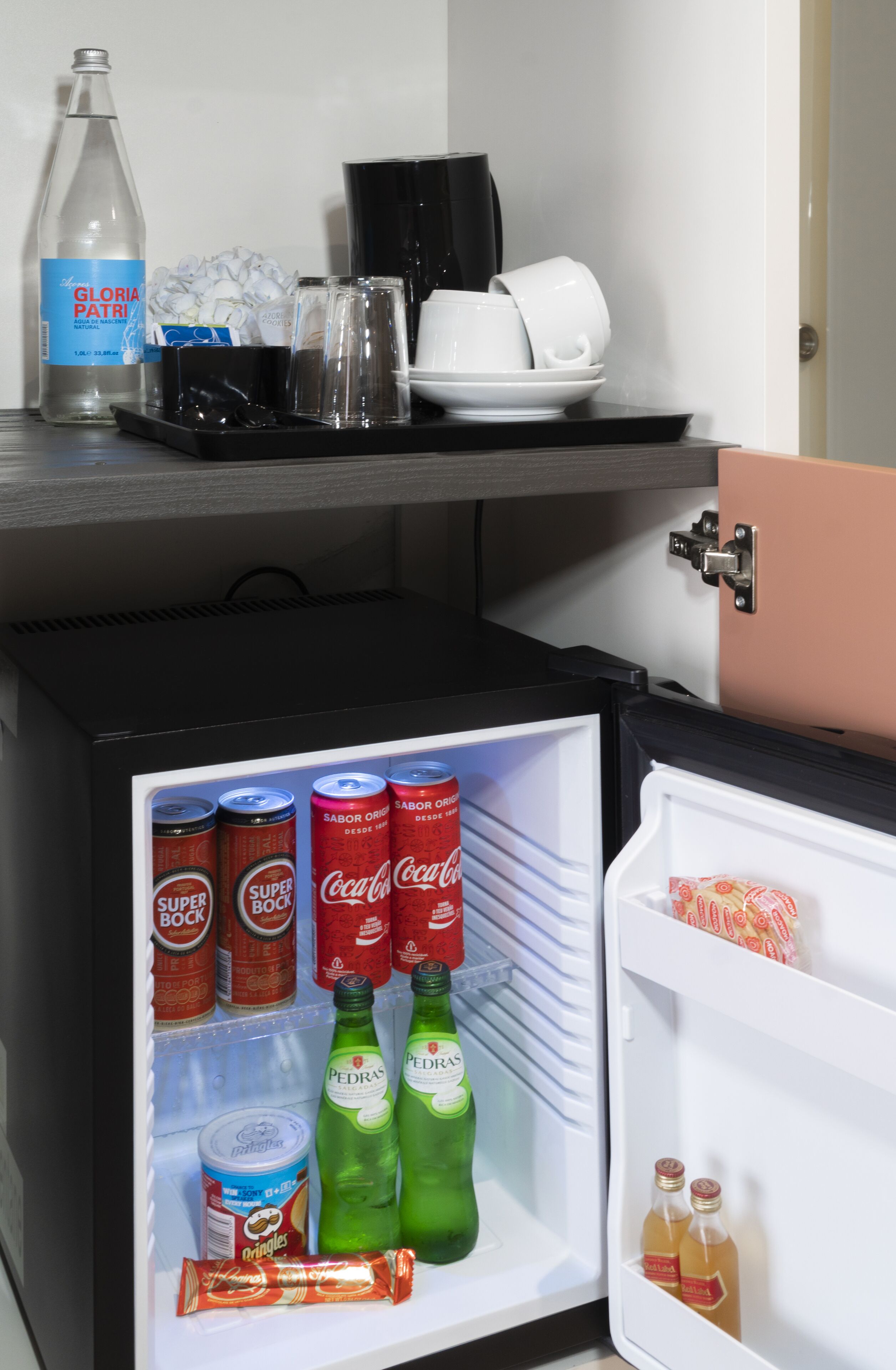 A mini-fridge stocked with beverages and snacks, a coffee maker, and a bottle of water on a countertop.