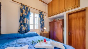 A bedroom with a blue bedspread, wooden wardrobe, and a window with curtains. - Casa La Perla (Rhodes)