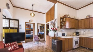 A spacious living room with a large flat-screen TV, a sofa with a 'LIVE LAUGH LOVE' cushion, a patterned rug, and a wooden dining table. The kitchen features wooden cabinets, a microwave, a stove, and a coffee maker. - Casa La Perla (Rhodes)