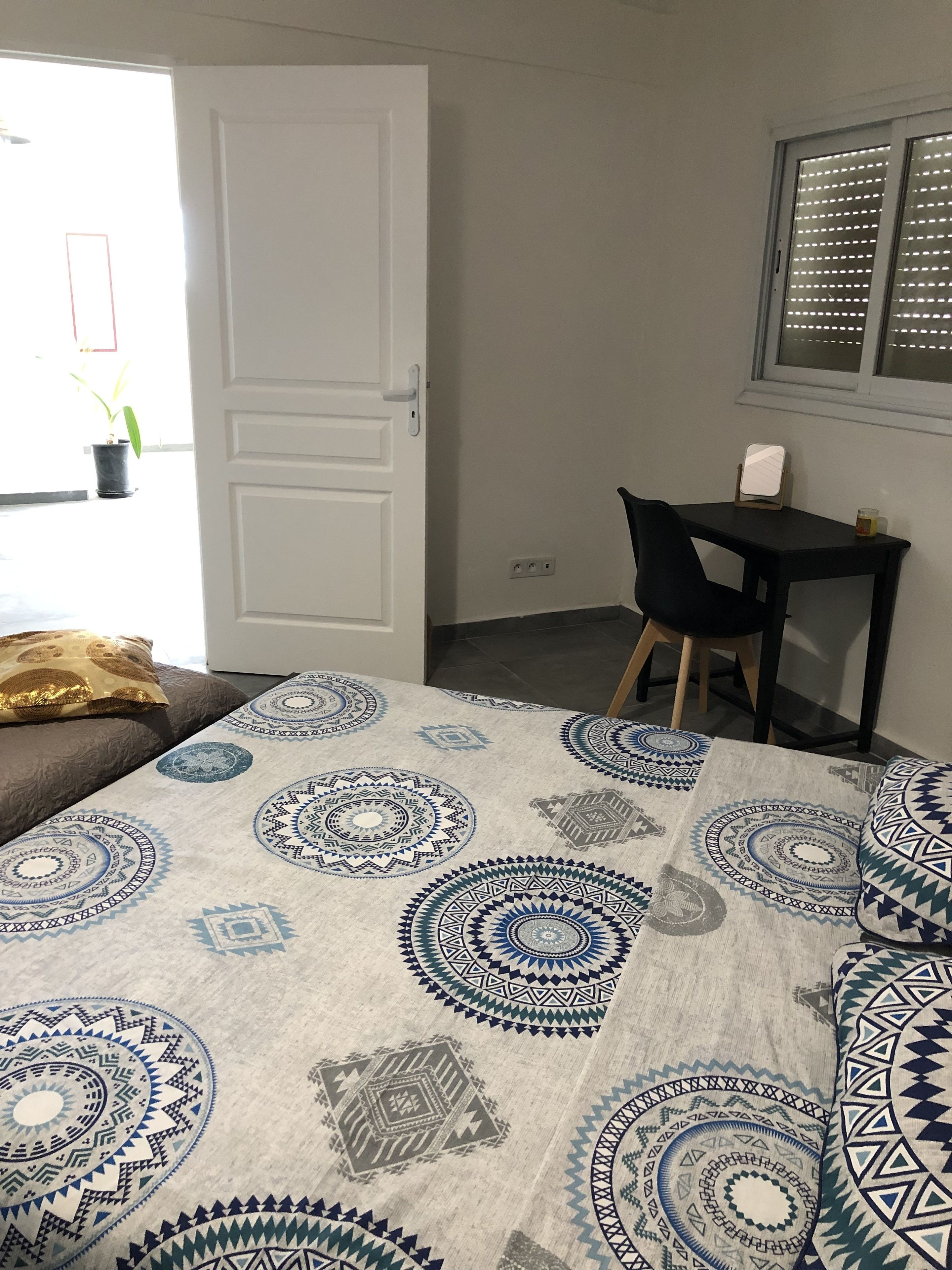 A bedroom with a bed covered in patterned bedding, a desk with a chair, a window with blinds, and a door leading outside.