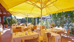 An outdoor dining area with yellow umbrellas, white tablecloths, wicker chairs, and a tiled floor. - Kamino (Häusern)