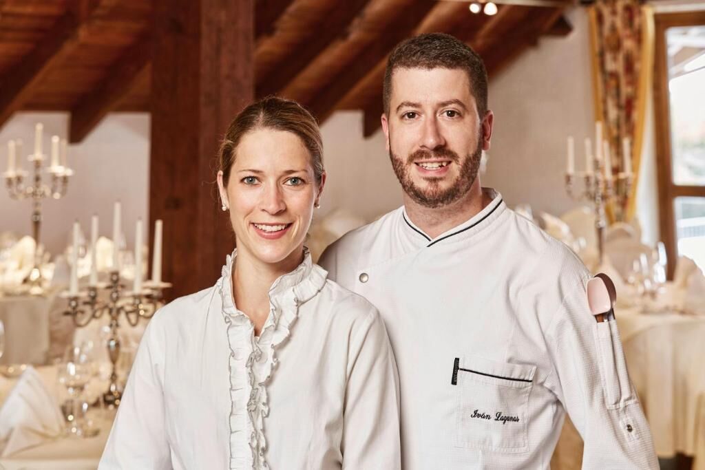 Dos personas con uniformes de chef blancos de pie en un comedor con una araña y mesas dispuestas.