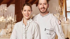 Two people in white chef uniforms standing in a dining area with a chandelier and table settings. - Kamino (Häusern)