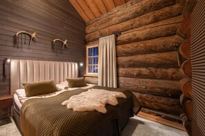 A log cabin bedroom with a bed, pillows, a fur throw, wooden walls, a window with curtains, and wall-mounted lights. - Kuukkeli Hirvas Suite (Saariselka)