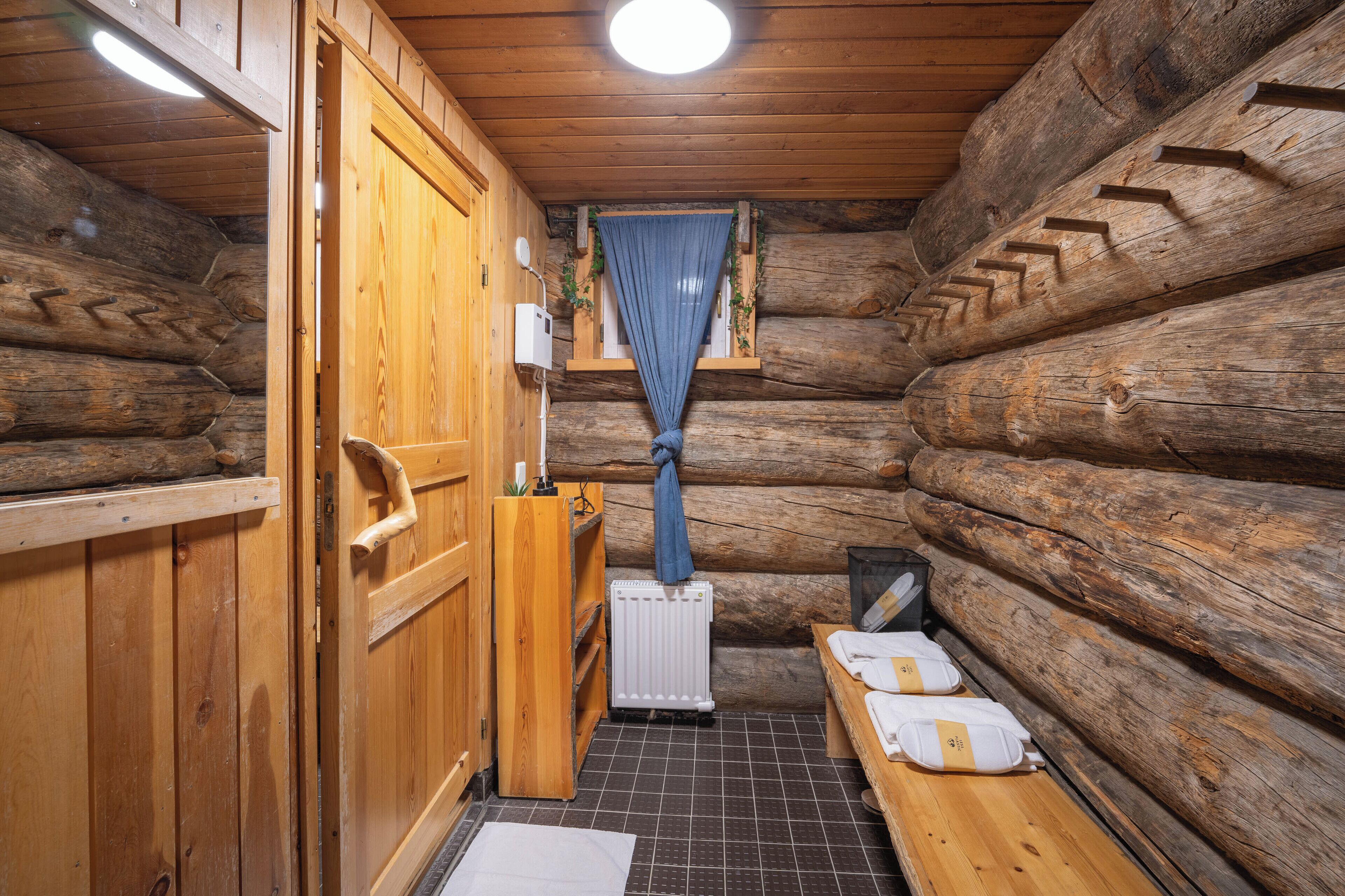 A log cabin bathroom with wooden walls, a wooden bench, a radiator, and a window with a blue curtain.