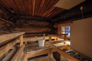 Interior de una cabaña de troncos con bancos de madera, una ventana y un balde blanco.