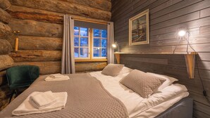 A log cabin room with a bed, a chair, a window with curtains, a painting, and a wall-mounted lamp. - Kuukkeli Hirvas Suite (Saariselka)