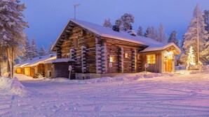 A log cabin with a snow-covered roof and a warm-lit entrance, surrounded by snow-covered trees. - Kuukkeli Hirvas Suite (Saariselka)