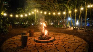 Uma fogueira em um padrão circular de pedra, rodeada por tocos de madeira, com luzes de corda acima.
