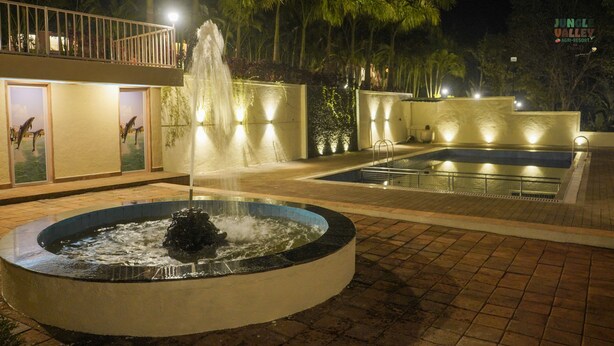 Um espelho de água circular com uma fonte no centro, rodeado por uma piscina e um caminho de azulejos.