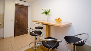 Een kleine kamer met een houten deur, een witte vaas met bloemen, een schaal met fruit en twee zwarte barkrukken.