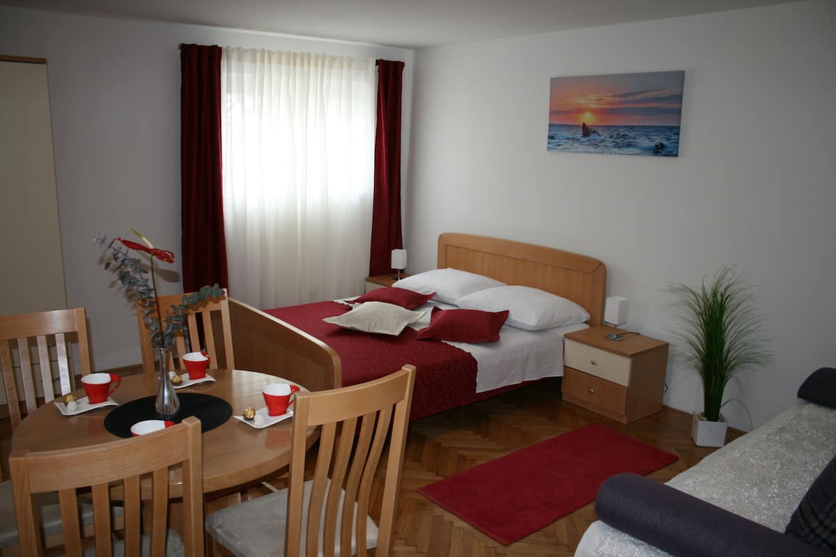 Una habitación de hotel con cama, una mesa de comedor con sillas, una mesita con tazas y un jarrón, y un cuadro de un atardecer en la pared.