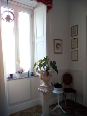 A room with a large window, a potted plant on a pedestal, a small table with a chair, and framed certificates on the wall.