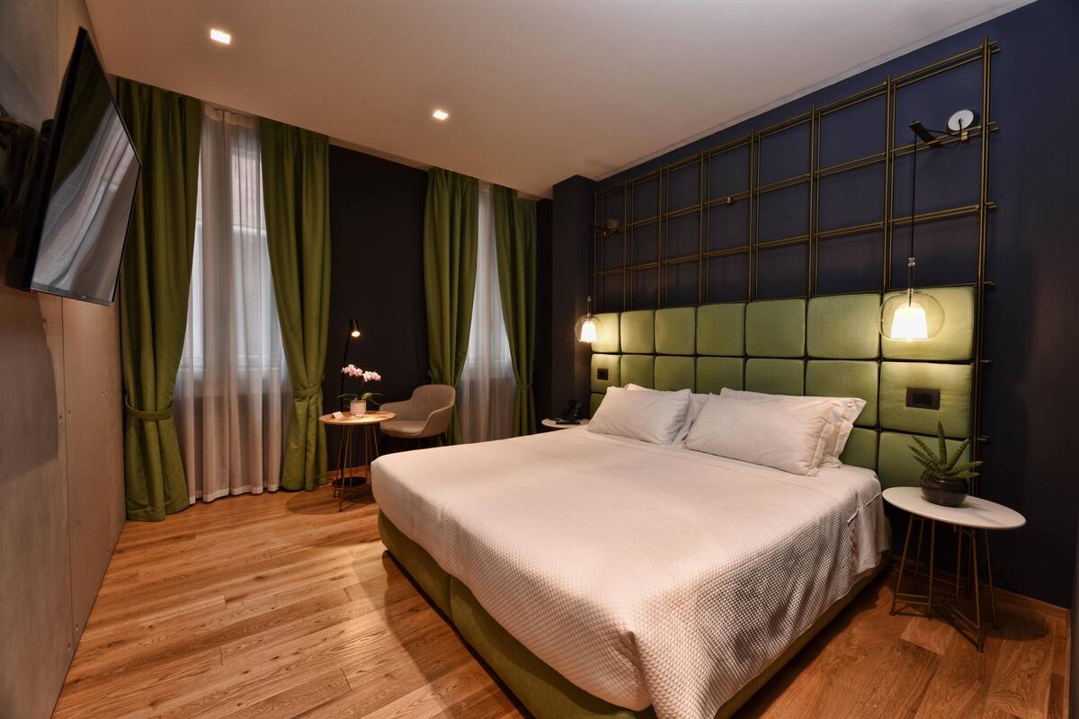A modern hotel room with a large bed, green curtains, a small round table, and a wall-mounted TV.
