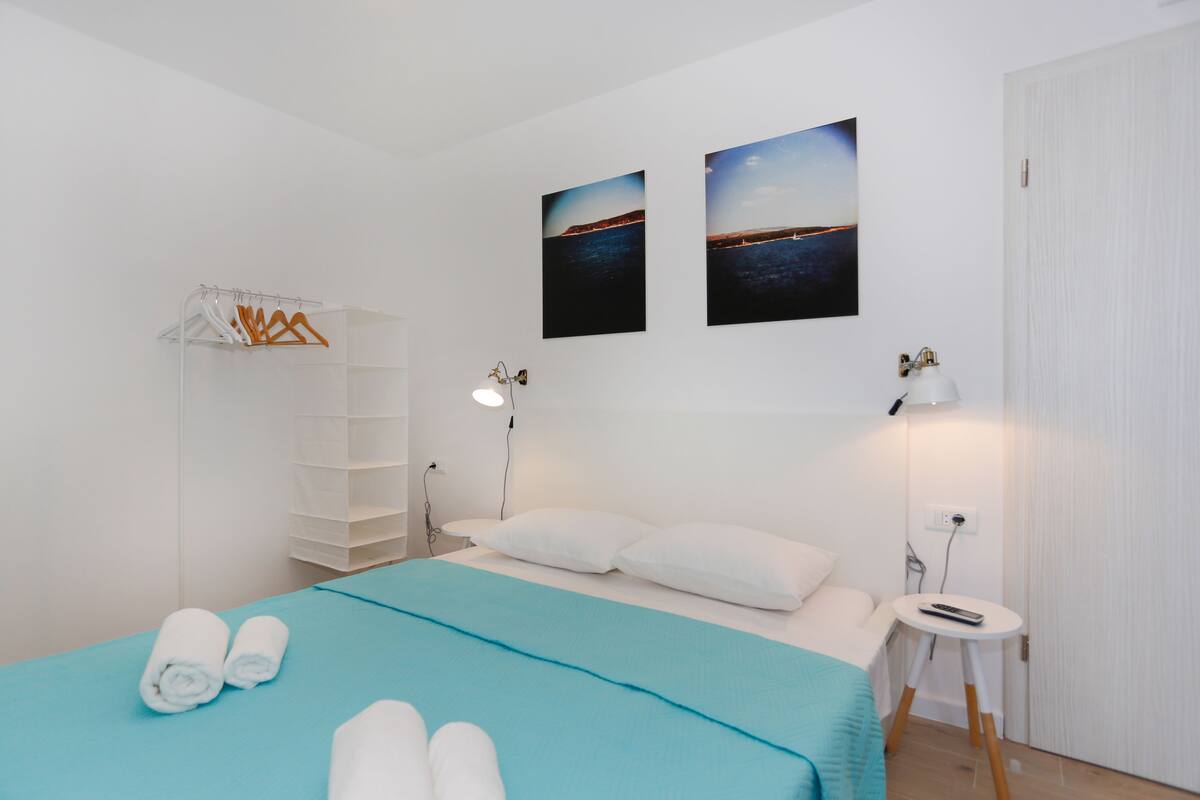 A modern hotel room with a bed, two bedside lamps, a small table, and a wall-mounted shelf.