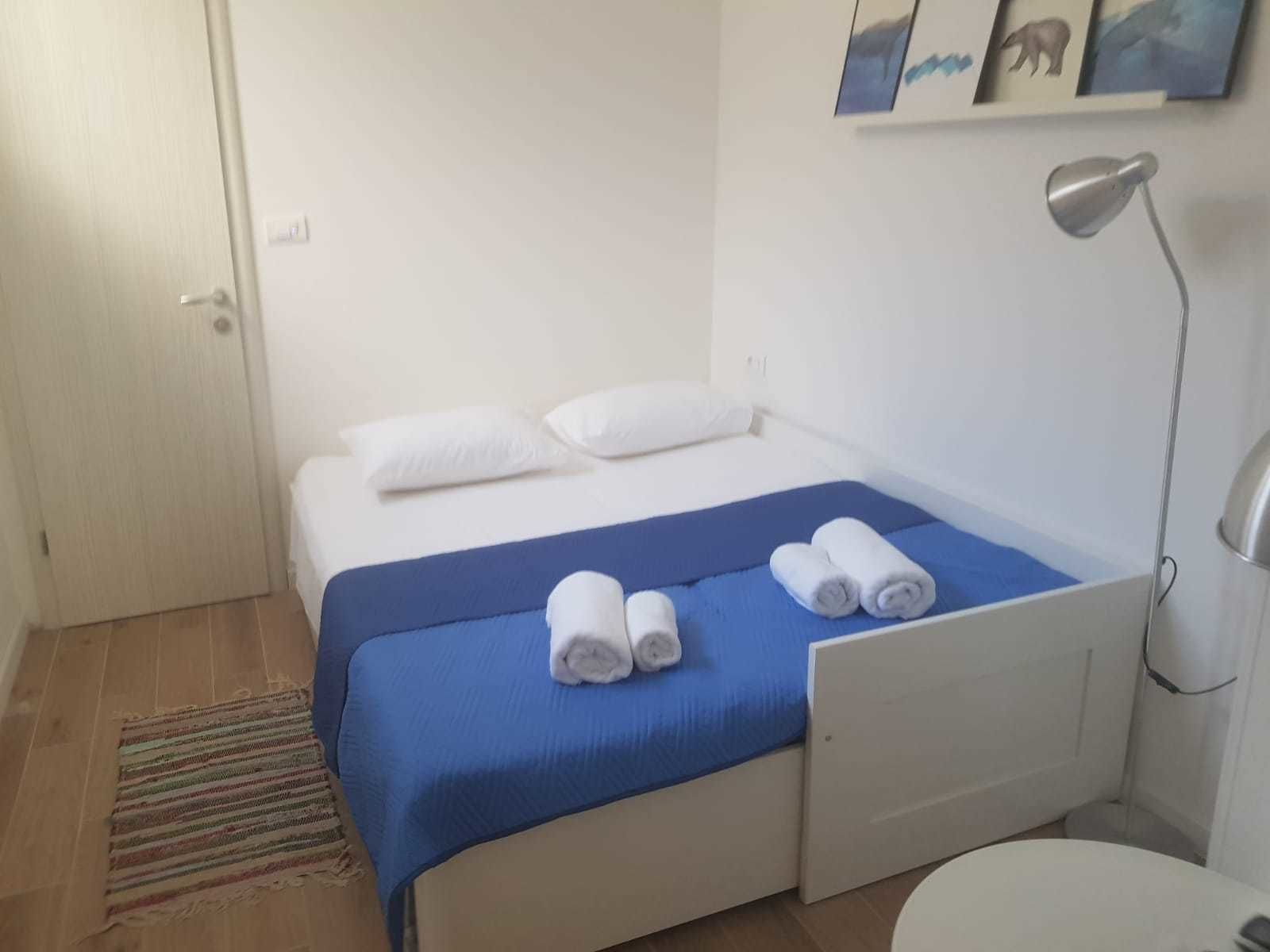 A small, clean room with a bed, a desk, and a chair. There are two rolled towels on the bed.