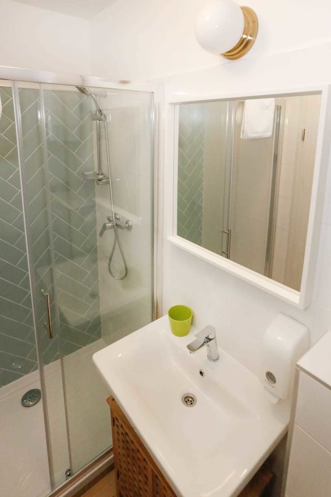 A bathroom with a glass-enclosed shower, a white sink with a green cup, and a mirror with a gold accent.