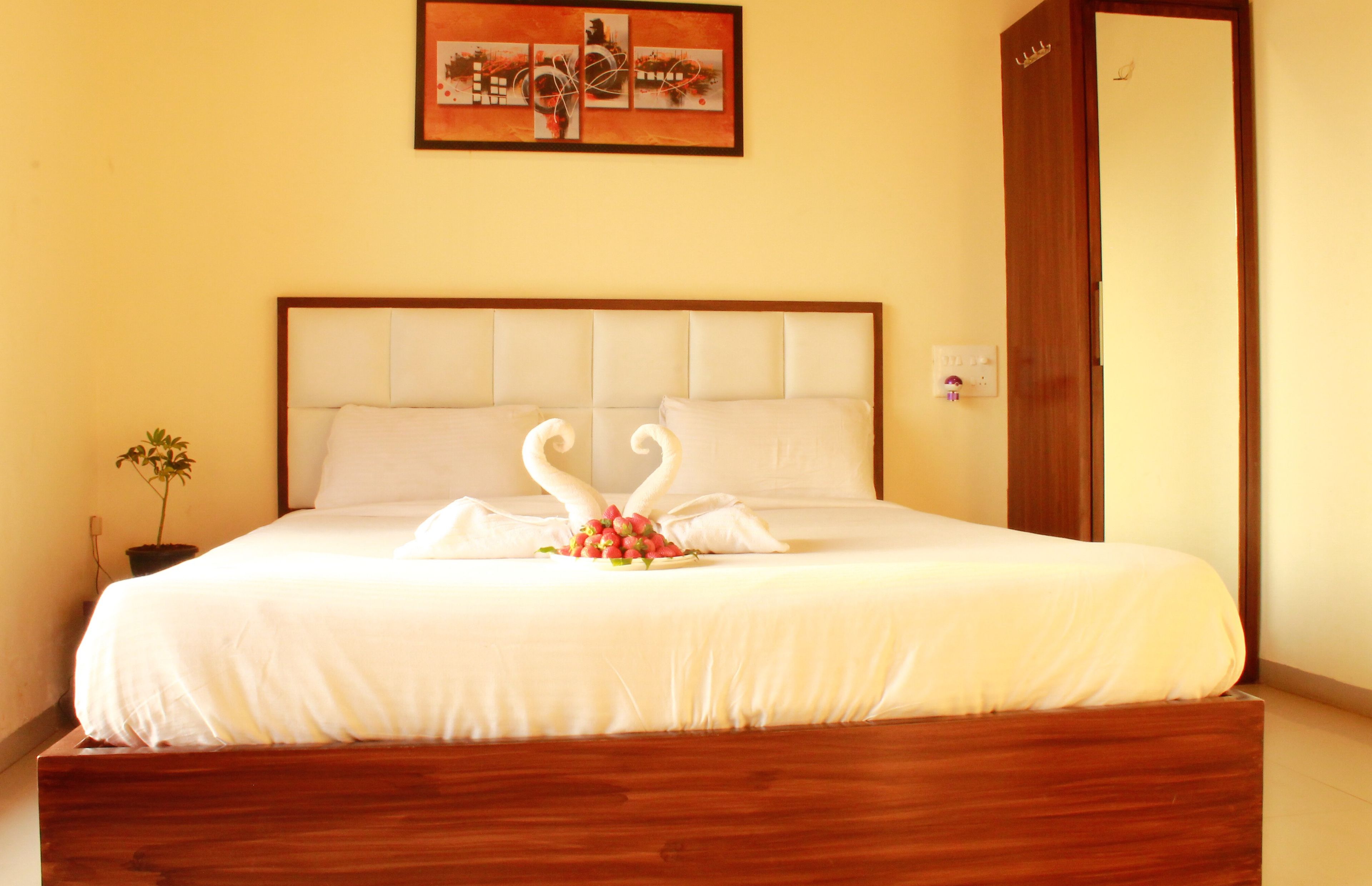 A neatly made bed with a white bedspread and two swan-shaped pillows, a small potted plant, and a painting on the wall.