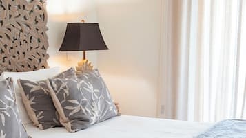 Una cama bien hecha con edredón blanco y almohadas grises, una lámpara oscura en una mesita de noche y un panel decorativo en la pared.
