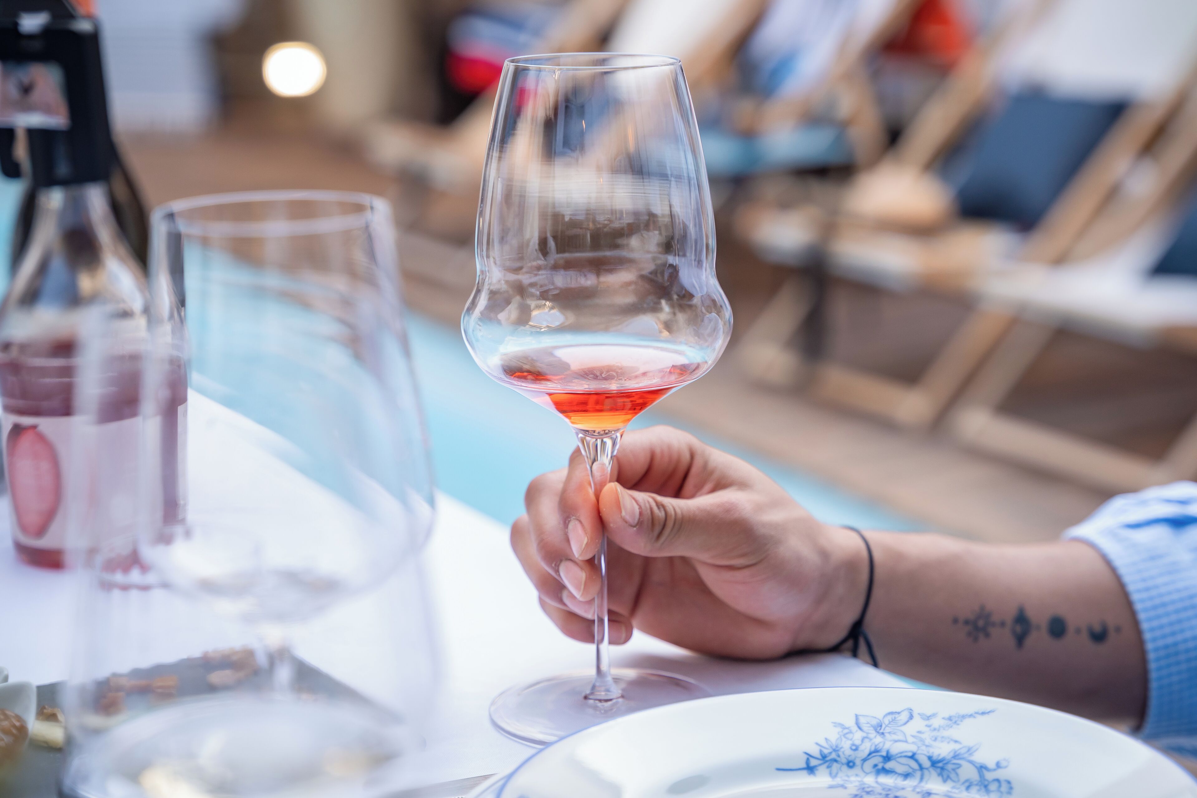 A person holding a wine glass with a red liquid, a bottle of wine, and a plate with a floral design.