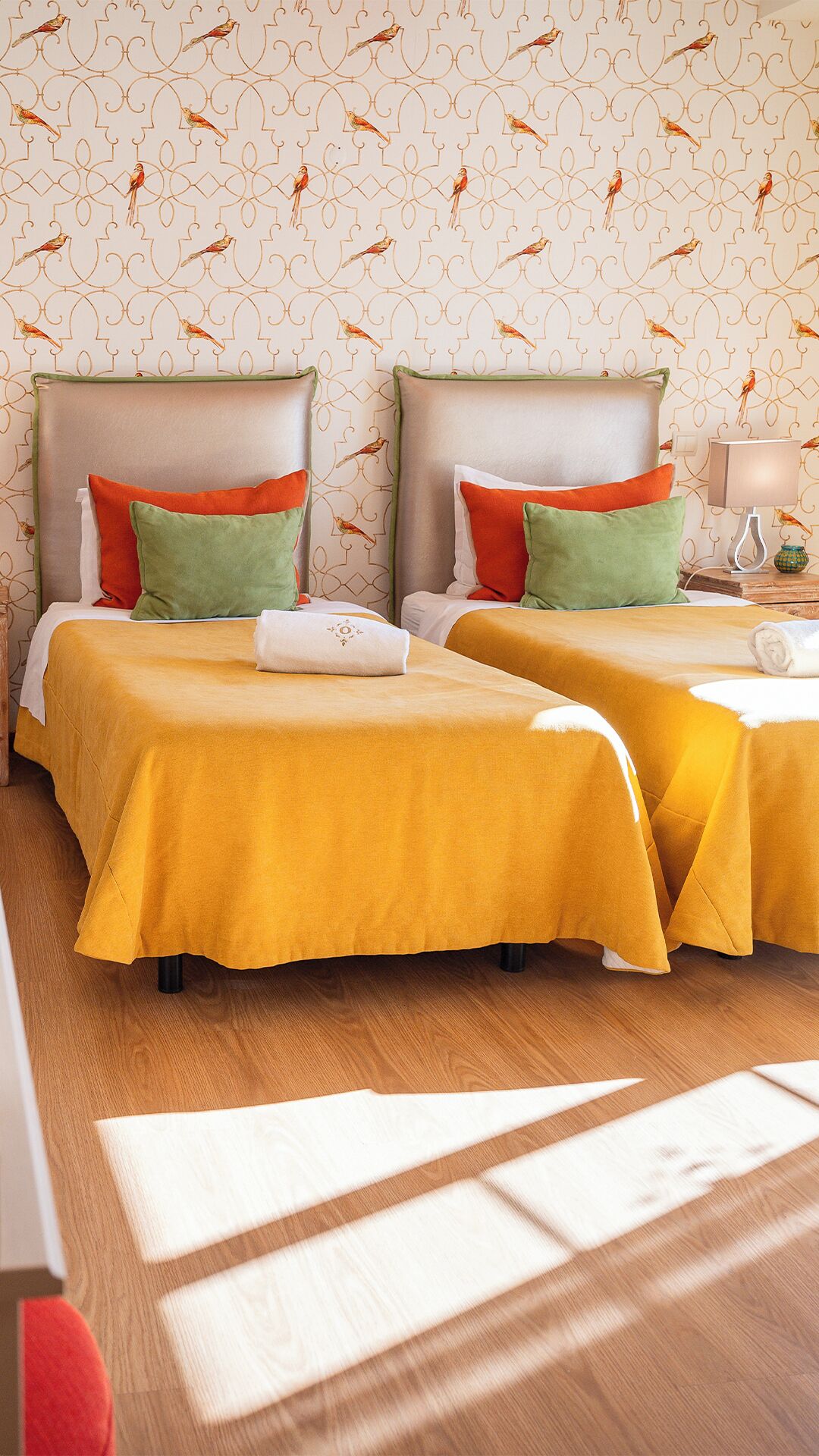 A hotel room with two beds, each with a yellow bedspread and pillows in red, green, and white. The room has a patterned wallpaper with birds and a wooden floor.